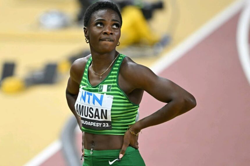 Female sprinter in a green striped uniform with bib reading AMUSAN and Budapest 23 standing with hands on hips on a track looking upward  trybalnews