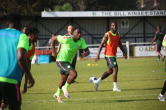 Football players in neon-green kits sprint during a training drill on a grassy pitch with a ball nearby.