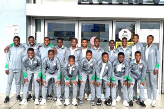 Group of 18 young athletes in matching gray tracksuits with green accents pose for a team photo outside a building.