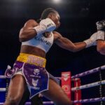Female boxer wearing a white sports bra and colorful shorts throws a punch in a boxing ring A gloved fist connects as the opponents arm extends into view  trybalnews