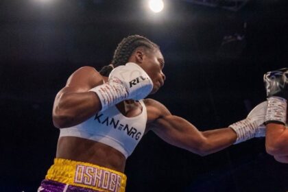 Female boxer wearing a white sports bra and colorful shorts throws a punch in a boxing ring. A gloved fist connects as the opponent’s arm extends into view.