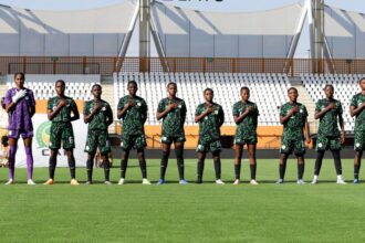 Soccer team lined up on a grassy field with hands over their hearts, wearing green uniforms in a stadium.”] ,