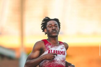 Male sprinter in Alabama red and white uniform, mid-race on a track with a focused expression.
