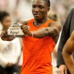Male sprinter in an orange sleeveless top holds up a race bib reading Kayinsola Ajayi during a track meet  trybalnews