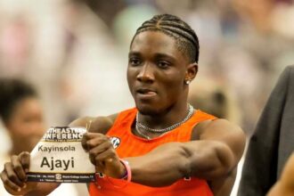 Male sprinter in an orange sleeveless top holds up a race bib reading 'Kayinsola Ajayi' during a track meet.