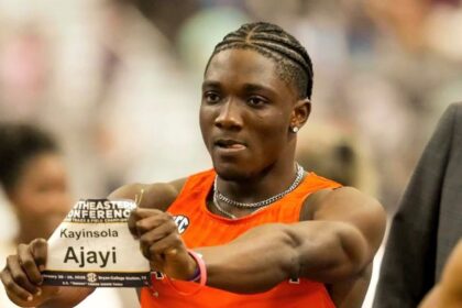 Male sprinter in an orange sleeveless top holds up a race bib reading 'Kayinsola Ajayi' during a track meet.