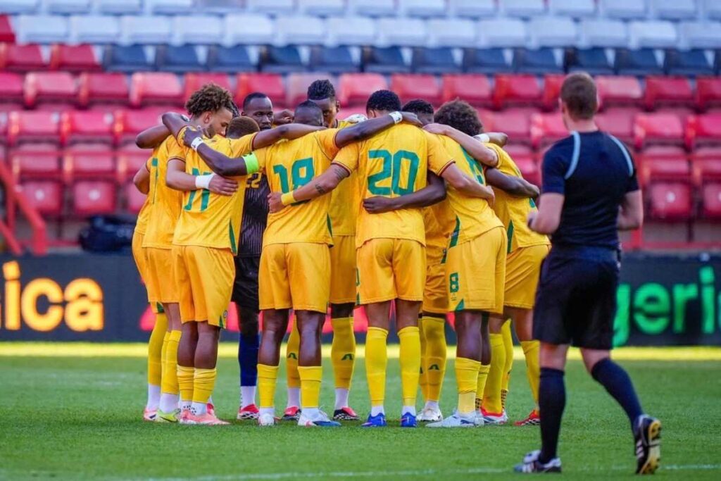 ZimbabweNationalTeam Trybal News Soccer team in yellow uniforms huddles together on the green field with a referee in black nearby and red stadium seats in the background trybalnews