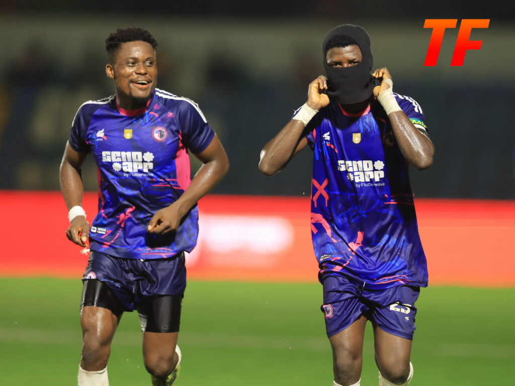 Two soccer players in blue kits sprint on a green field one wearing a black balaclava mask on his head  trybalnews
