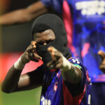 Athlete wearing a black balaclava and blue jersey points toward the camera with a colorful stadium backdrop behind them  trybalnews