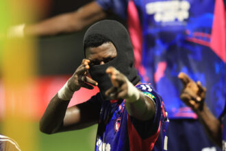 Athlete wearing a black balaclava and blue jersey points toward the camera with a colorful stadium backdrop behind them.