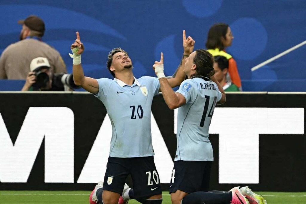 Uruguay midfielder Agustin Canobbio wearing number 14 light blue jersey with dark shorts kneels on the pitch pointing upward with his right index finger Teammate Maximiliano Araujo 20 in matching light blue jersey stands beside him with arms spread wide and head tilted back celebrating Both players have white wrist tape Stadium advertising boards and blurred crowd in background at Miami Stadium June 21 2026 during FIFA World Cup 2026 Group H match vs Cabo Verde  trybalnews