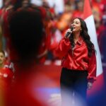Female singer wearing a bright red jacket performs into a handheld microphone on a crowded stage  trybalnews