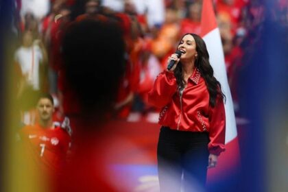 Female singer wearing a bright red jacket performs into a handheld microphone on a crowded stage.