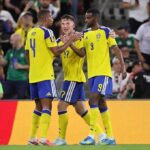 Three Sweden soccer players in yellow jerseys celebrate together on the field during a match with a cheering crowd behind them  trybalnews