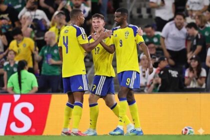 Three Sweden soccer players in yellow jerseys celebrate together on the field during a match, with a cheering crowd behind them.