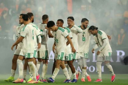 Soccer players in white and green kits huddle together on the field after a match, with colorful cleats and a smoky stadium in the background.