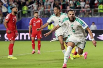 Amine Gouiri in Algeria’s white #9 jersey with green stripes runs forward with mouth open and arms outstretched in celebration. He wears white shorts, socks with "FAF", and pink football boots. Behind him, an Algeria teammate in the same kit runs and shouts with his right fist clenched. Two Jordan players in red #5 and #11 jerseys stand dejected in the background. The match is Jordan vs Algeria at San Francisco Bay Area Stadium, FIFA World Cup 2026 Group J, June 22 2026, Santa Clara California. A packed stadium crowd is blurred behind a blue advertising board.