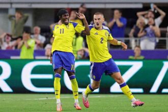 Sweden forward Anthony Elanga wearing yellow #11 jersey with blue shoulders, blue shorts, yellow socks, and a black headband celebrates with teammate Gustaf Lagerbielke #2. Elanga is on the left with his mouth open shouting, right arm raised, and Lagerbielke’s arm around his shoulder. Lagerbielke, wearing yellow #2 jersey and blue shorts, runs beside him with his mouth open and left arm out. Both wear matching yellow Adidas kits with Sweden crest and blue shorts. FIFA World Cup 2026 Group F match between Japan and Sweden at Dallas Stadium, Arlington, Texas on June 25, 2026. Blurred fans react in the background.
