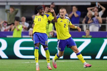 Sweden forward Anthony Elanga wearing yellow #11 jersey with blue shoulders, blue shorts, yellow socks, and a black headband celebrates with teammate Gustaf Lagerbielke #2. Elanga is on the left with his mouth open shouting, right arm raised, and Lagerbielke’s arm around his shoulder. Lagerbielke, wearing yellow #2 jersey and blue shorts, runs beside him with his mouth open and left arm out. Both wear matching yellow Adidas kits with Sweden crest and blue shorts. FIFA World Cup 2026 Group F match between Japan and Sweden at Dallas Stadium, Arlington, Texas on June 25, 2026. Blurred fans react in the background.