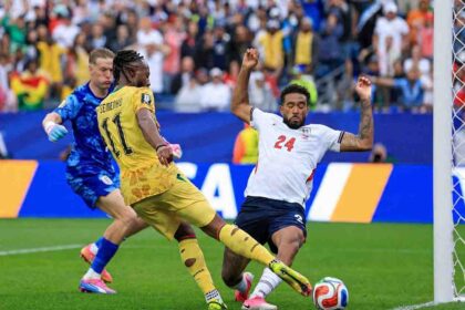 Ghana forward Antoine Semenyo wearing yellow #11 jersey attempts a shot on goal as England defender Reece James in white #24 jersey blocks the attempt. Action near goalpost during FIFA World Cup 2026 Group L match between England and Ghana at Boston Stadium, Foxborough Massachusetts on June 23, 2026. Goalkeeper visible in background.