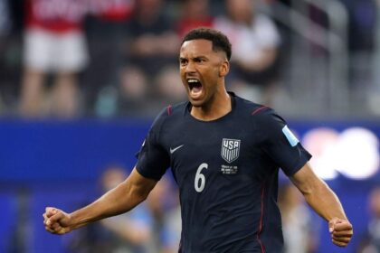 USA defender Auston Trusty wearing navy blue #6 Nike jersey with white USA crest and red trim celebrates with a roar. He has short curly hair, both fists clenched, and arms spread wide with an intense expression. FIFA World Cup 2026 Group D match between Türkiye and USA at Los Angeles Stadium, Inglewood, California on June 25, 2026. Blurred crowd and blue stadium boards in the background.