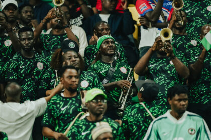Crowd of band members in green patterned jerseys playing brass instruments at a stadium-like event.