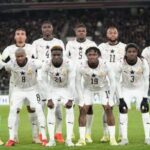 Eleven soccer players in white uniforms for Ghana pose for a team photo on the field numbers visible on jerseys stadium crowd in the background  trybalnews