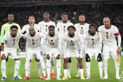 Eleven soccer players in white uniforms for Ghana pose for a team photo on the field, numbers visible on jerseys, stadium crowd in the background.