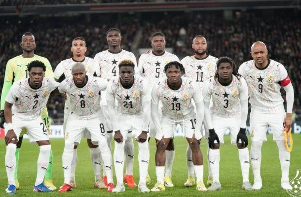 Eleven soccer players in white uniforms for Ghana pose for a team photo on the field numbers visible on jerseys stadium crowd in the background  trybalnews