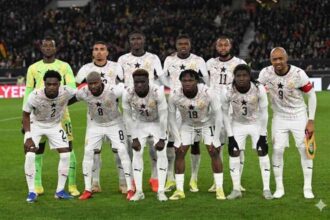 Ghana football team in white kits with star crests posing for a group photo on the pitch before a match, with fans in the stands behind them.