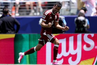 Male soccer player in a maroon jersey (#16) celebrating with clenched fists on the pitch.