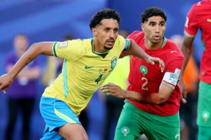 Two soccer players, one in yellow Brazil kit and one in red Morocco kit, chase the ball in a crowded match.