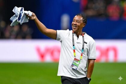 Sports official on a field, smiling and waving a folded shirt above his head.
