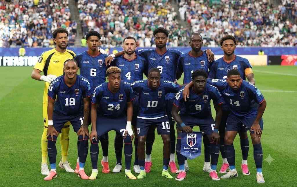 Cabo Verde National Team Trybal News Cabo Verde national football team pose for an official team photograph on the pitch before kickoff Eleven players in two rows wear dark blue home kits with red trim white numbers and the FCF crest The goalkeeper on the far left wears a yellow kit with white gloves Players 19 24 10 8 14 are crouching in the front row Players 6 4 3 are in the back row They stand on green grass with a blue trybalnews"CABO VERDE" pennant in front. 2026 FIFA World Cup Group H match vs Saudi Arabia on June 26, 2026 at Houston Stadium in Houston, Texas. A packed stadium crowd is blurred in the background.