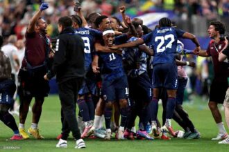 Cabo Verde players in dark blue kits with white numbers celebrate on the pitch after a 0-0 draw. Player #11 wearing a white headband smiles and is hugged in the center, with teammate #22 "MOREIRA" to his right and #9 "MENDES" behind him. A staff member in a maroon shirt raises a blue-gloved fist on the left, and another staff member holds a camera on the right. 2026 FIFA World Cup Group H match between Cabo Verde and Saudi Arabia on June 26, 2026 at Houston Stadium in Houston, Texas. A blurred crowd fills the stadium background.