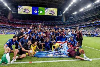 Cabo Verde football players and coaching staff in blue and yellow kits, plus black and maroon staff shirts, pose together on the pitch celebrating. They are gathered around a large blue banner with “FORÇA CABO VERDE” and a shark graphic, and one player holds the Cabo Verde national flag. The scoreboard above shows 0-0. 2026 FIFA World Cup match between Cabo Verde and Saudi Arabia on June 26, 2026 at Houston Stadium in Houston, United States. A packed stadium with blue and red seats is in the background.