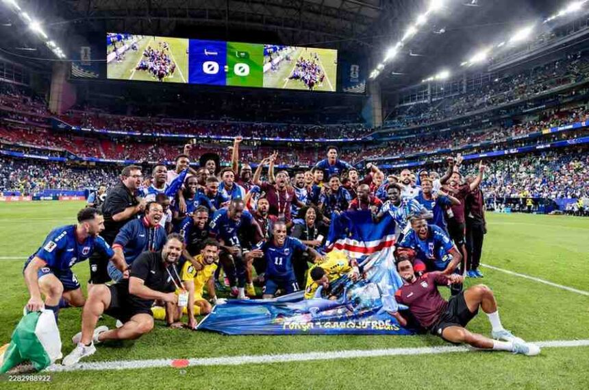 Cabo Verde football players and coaching staff in blue and yellow kits plus black and maroon staff shirts pose together on the pitch celebrating They are gathered around a large blue banner with FORÇA CABO VERDE and a shark graphic and one player holds the Cabo Verde national flag The scoreboard above shows 0 0 2026 FIFA World Cup match between Cabo Verde and Saudi Arabia on June 26 2026 at Houston Stadium in Houston United States A packed stadium with blue and red seats is in the background  trybalnews