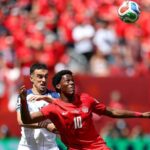 Two soccer players compete for the ball during a match the player in red Canada 10 ahead of a white jersey opponent as the ball flies overhead with a red clad crowd in the background  trybalnews