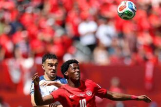 Two soccer players compete for the ball during a match, the player in red (Canada, #10) ahead of a white-jersey opponent as the ball flies overhead with a red-clad crowd in the background.