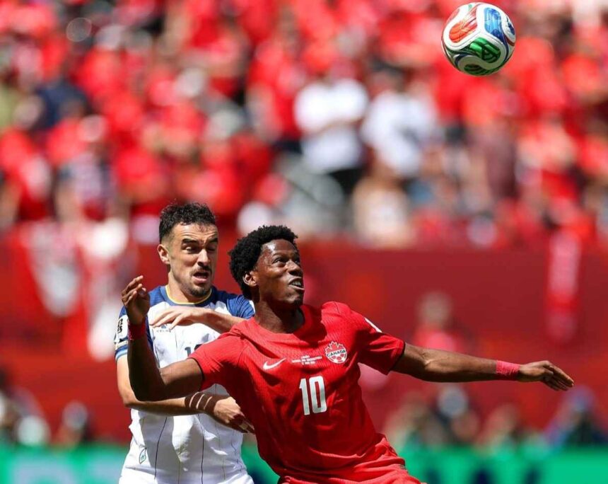 Two soccer players compete for the ball during a match the player in red Canada 10 ahead of a white jersey opponent as the ball flies overhead with a red clad crowd in the background  trybalnews
