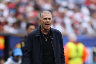 Ghana head coach Carlos Queiroz stands on the sideline wearing a navy jacket and black shirt with a gold necklace. He appears to be shouting instructions with an intense expression during the FIFA World Cup 2026 Group L match between England and Ghana at Boston Stadium, Foxborough Massachusetts on June 23, 2026. Blurred crowd and stadium staff visible in background.