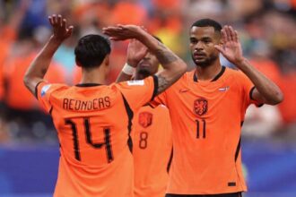 Netherlands players Cody Gakpo, wearing orange jersey number 11, and Tijjani Reijnders, wearing orange jersey number 14, celebrate together on the pitch. Gakpo faces the camera with both hands raised, while Reijnders has his back to camera with arms up high. Both wear orange Netherlands kits with the Dutch crest. A third teammate in jersey number 8 is blurred in the background. Fans in orange fill the stadium stands behind them. Match was FIFA World Cup 2026 Group F, Netherlands vs Sweden at Houston Stadium, Houston, Texas on June 20, 2026.