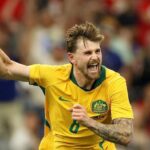Australian soccer player in a yellow jersey celebrates a goal fist raised and shouting  trybalnews