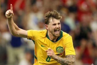 Australian soccer player in a yellow jersey celebrates a goal, fist raised and shouting