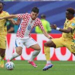 Croatia midfielder Martin Baturina wearing a red and white checkered kit with number 16 controls the ball in the center He is challenged by two Ghana players in yellow kits Marvin Senaya number 11 on the left and Ghana 26 on the right The 2026 FIFA World Cup Group L match between Croatia and Ghana took place on June 27 2026 at Philadelphia Stadium in Philadelphia Pennsylvania Stadium advertising boards and a green pitch are visible in the background  trybalnews