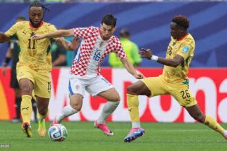 Croatia midfielder Martin Baturina, wearing a red and white checkered kit with number 16, controls the ball in the center. He is challenged by two Ghana players in yellow kits: Marvin Senaya, number 11 on the left, and Ghana #26 on the right. The 2026 FIFA World Cup Group L match between Croatia and Ghana took place on June 27, 2026 at Philadelphia Stadium in Philadelphia, Pennsylvania. Stadium advertising boards and a green pitch are visible in the background.