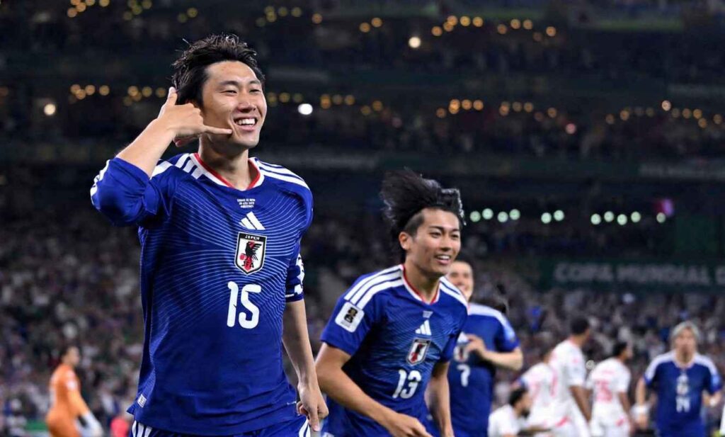 Japan midfielder Daichi Kamada wearing the blue Japan 15 jersey with white Adidas stripes and the JFA crest celebrates scoring Japans first goal He smiles broadly and makes a call me hand gesture near his face with his right hand Behind him Japan teammate 13 runs toward him smiling with other players and a packed stadium blurred in the background under stadium lights The match was FIFA World Cup 2026 Group F Tunisia vs Japan at Monterrey Stadium Meixco  trybalnews
