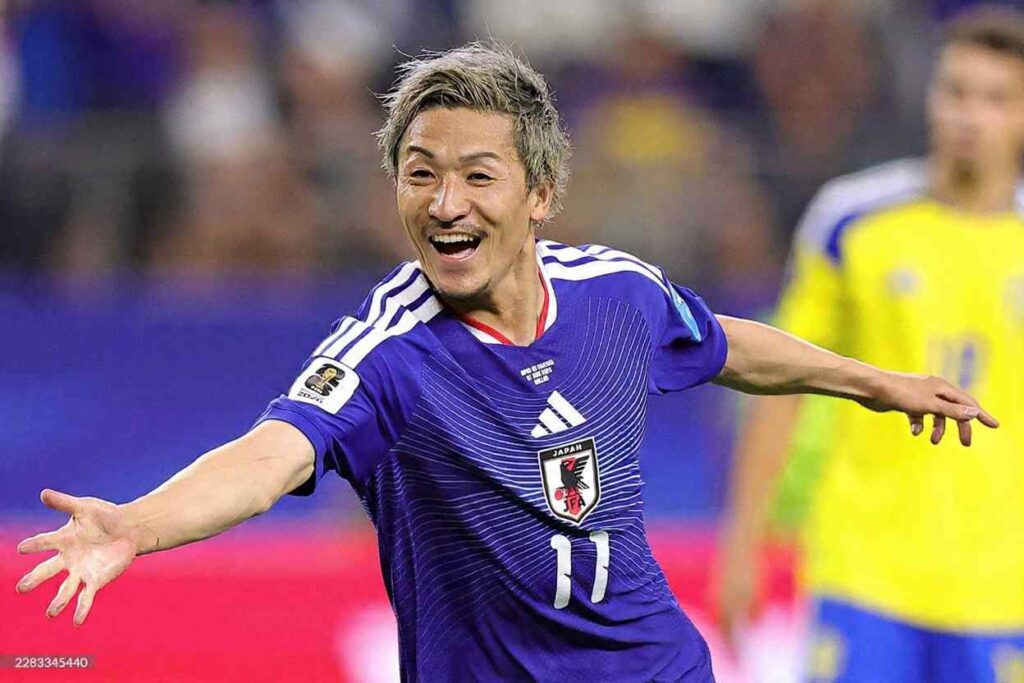 Japan forward Daizen Maeda wearing blue 11 Adidas jersey with white shoulder stripes red collar trim and Japan Football Association crest celebrates with arms outstretched and a wide smile He has bleached blonde hair with darker roots and light facial hair FIFA World Cup 2026 Group F match between Japan and Sweden at Dallas Stadium Arlington Texas on June 25 2026 A blurred Sweden player in yellow 11 kit is visible in the background  trybalnews