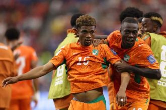 Cote d’Ivoire midfielder Amad Diallo, wearing an orange #15 Puma kit with green accents and curly brown hair, celebrates with his mouth open and arm outstretched. Teammates in matching orange kits, including #20, surround him with big smiles after scoring Cote d’Ivoire’s first goal. This is during the FIFA World Cup 2026 Round of 32 match between Cote d’Ivoire and Norway on June 30, 2026 at Dallas Stadium in Arlington, Texas. The background shows a blurred crowd and a player in a green training bib.