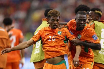 Cote d’Ivoire midfielder Amad Diallo, wearing an orange #15 Puma kit with green accents and curly brown hair, celebrates with his mouth open and arm outstretched. Teammates in matching orange kits, including #20, surround him with big smiles after scoring Cote d’Ivoire’s first goal. This is during the FIFA World Cup 2026 Round of 32 match between Cote d’Ivoire and Norway on June 30, 2026 at Dallas Stadium in Arlington, Texas. The background shows a blurred crowd and a player in a green training bib.